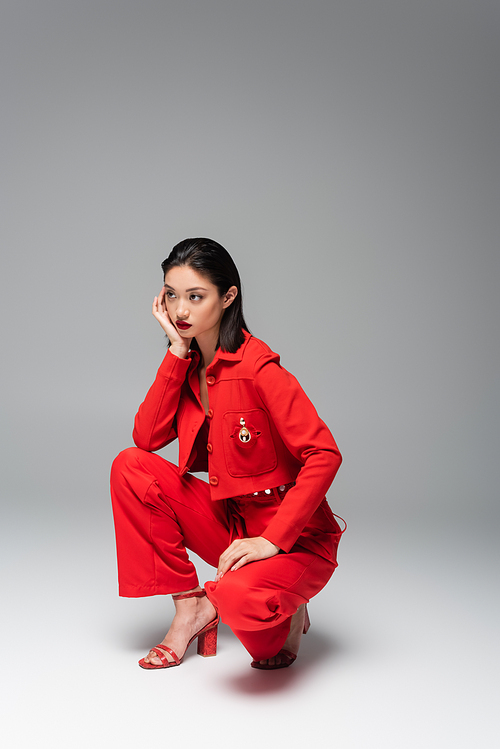 full length of asian woman in red and stylish outfit sitting on haunches with hand near face on grey background
