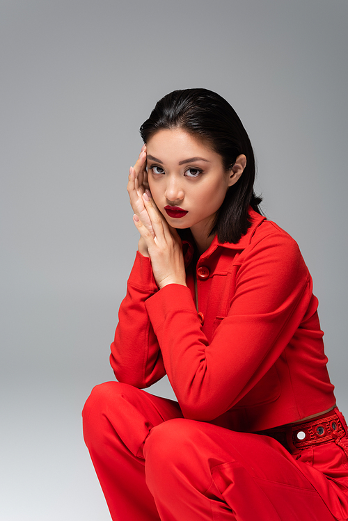 stylish asian woman in red elegant attire sitting on haunches with hands near face isolated on grey