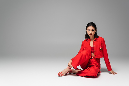 brunette asian woman in red trousers and jacket sitting on grey background