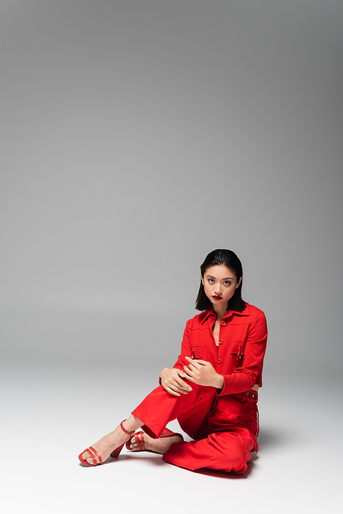 brunette asian model in red jacket and pants looking at camera while sitting on grey background