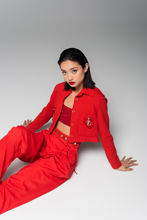 young asian woman in red fashionable suit looking at camera and sitting on grey background