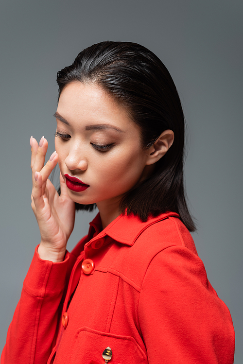 young asian woman in elegant jacket touching face with makeup and red lips isolated on grey
