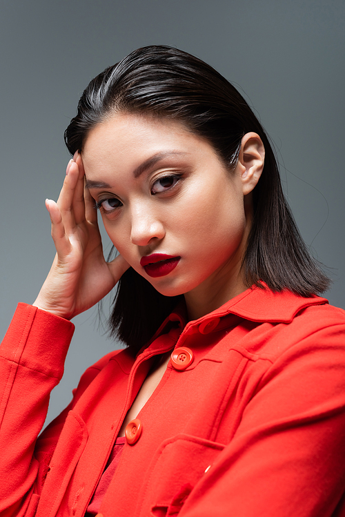 portrait of asian woman with makeup and red lips wearing fashionable jacket isolated on grey