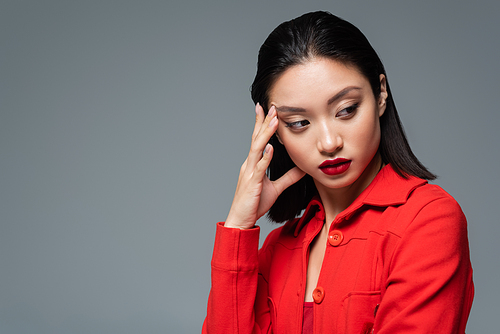 portrait of brunette asian woman in red jacket holding hand near face and looking away isolated on grey