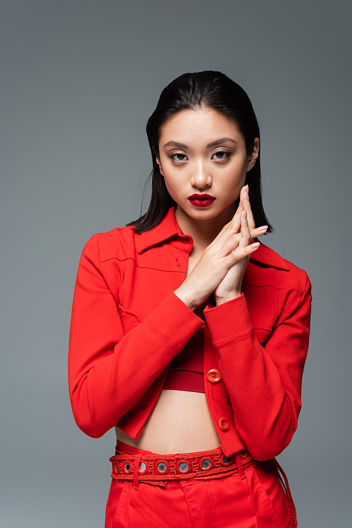 brunette asian model in red fashionable jacket looking at camera isolated on grey