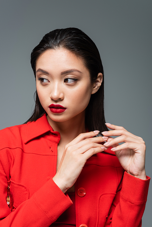 pretty asian woman with makeup and red lips wearing trendy jacket and looking away isolated on grey