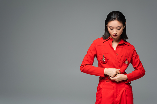 brunette asian woman touching red jacket decorated with brooches isolated on grey