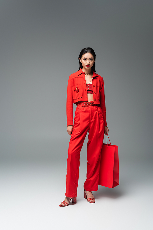 full length of asian model in red jacket and trousers standing with shopping bag on grey background