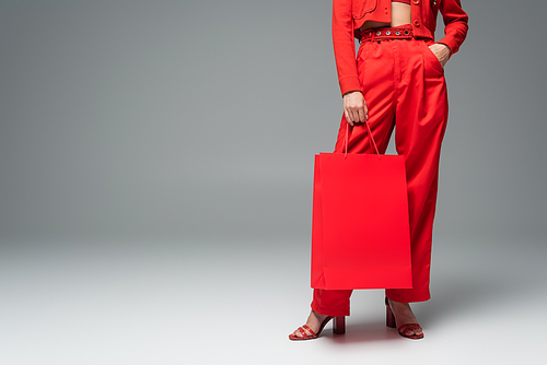 cropped view of woman holding shopping bag while standing with hand in pocket of red pants on grey background