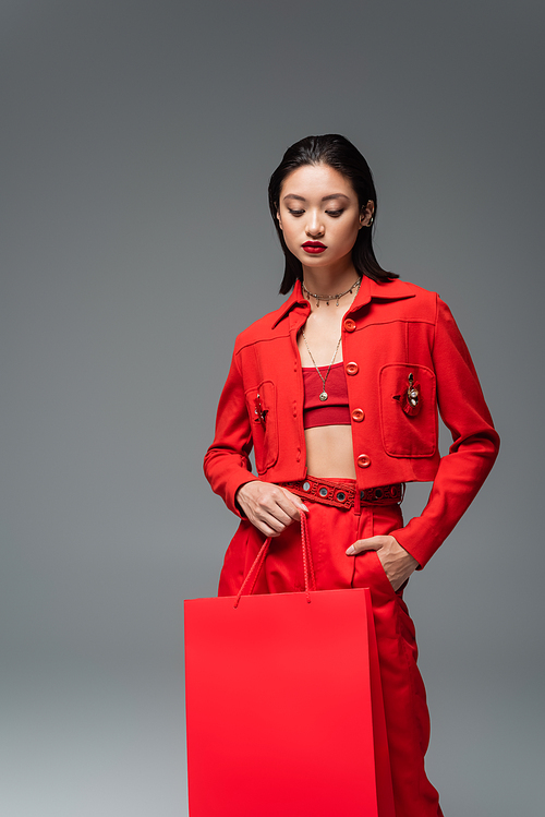 brunette asian woman in red stylish attire holding shopping bag and hand in pocket isolated on grey