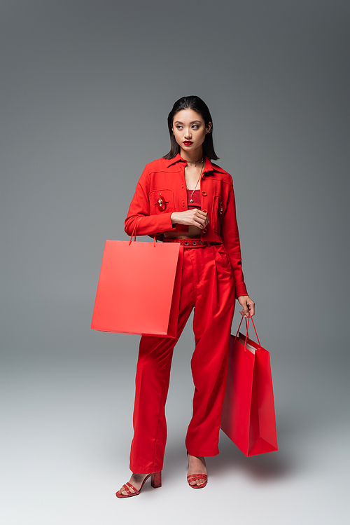 full length of asian woman in stylish jacket and pants holding shopping bags while looking away on grey background