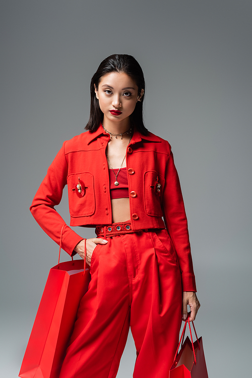 brunette asian woman in red fashionable clothes posing with hand in pocket and shopping bags isolated on grey