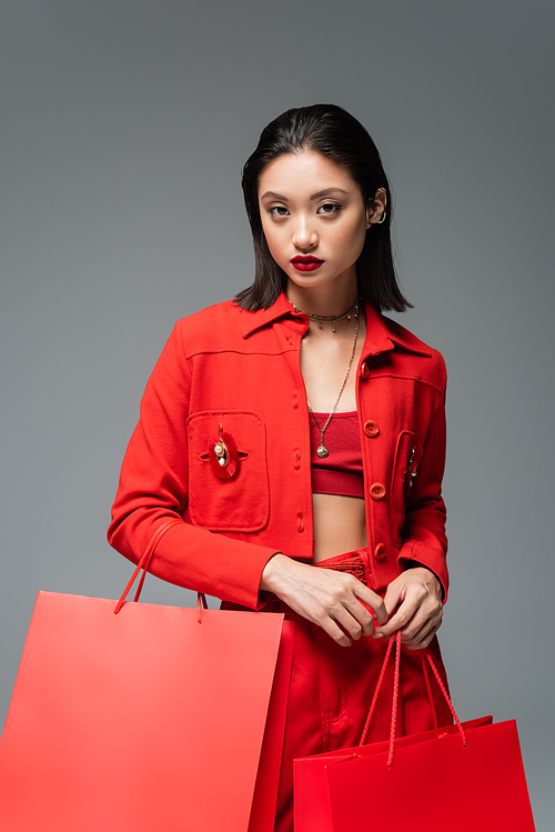 fashionable asian woman in red jacket holding shopping bags and looking at camera isolated on grey