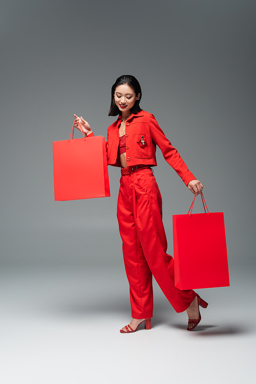 full length of smiling asian woman in red suit and heeled sandals posing with shopping bags on grey background