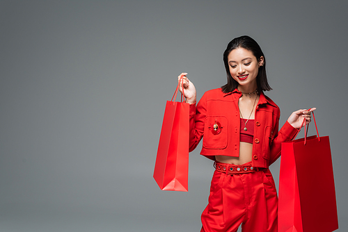 happy asian woman in elegant clothes posing with red shopping bags isolated on grey