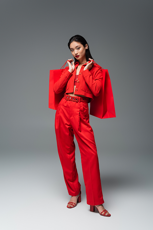 full length of asian woman in red outfit holding shopping bags and looking at camera on grey background