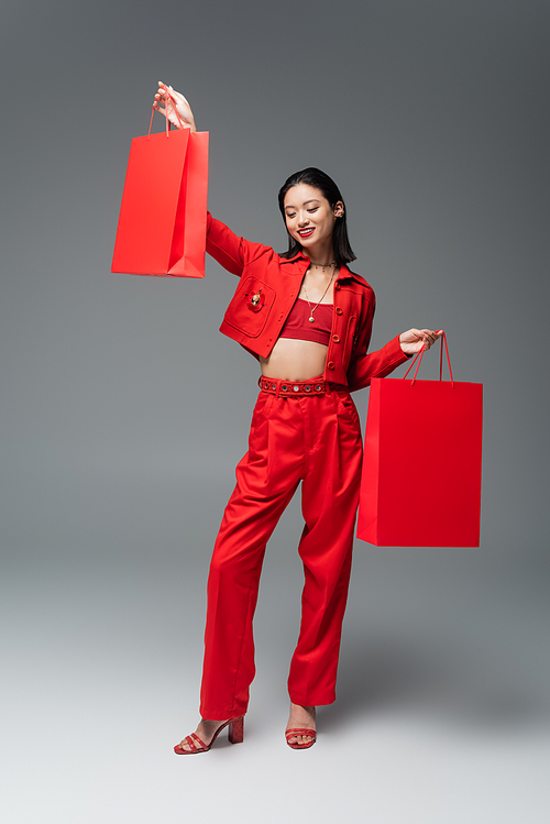 full length of cheerful asian woman in trendy attire posing with red shopping bags on grey background