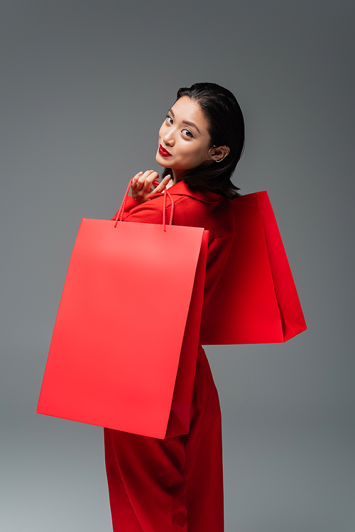 brunette asian woman with red shopping bags smiling at camera isolated on grey