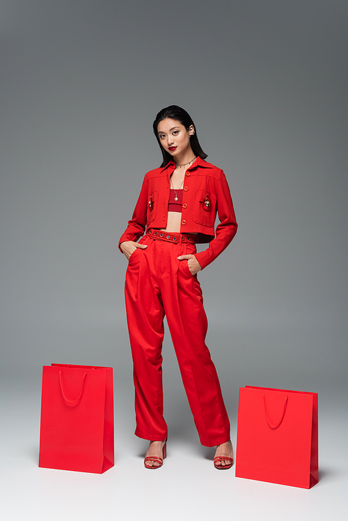 young asian model in red jacket and trousers posing with hands in pockets near shopping bags on grey background