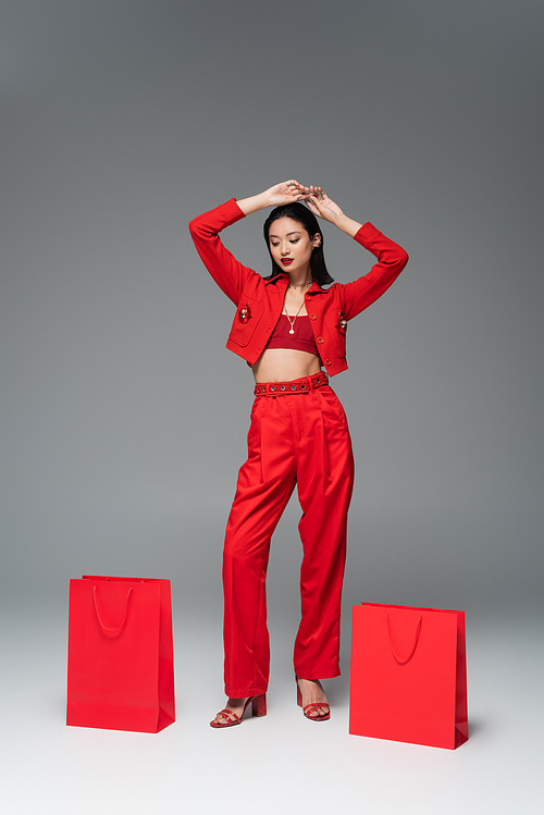 full length of asian woman in stylish outfit posing with hands above head near red shopping bags on grey background