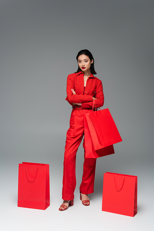 full length of asian woman in red stylish suit standing with crossed arms near shopping bags on grey background