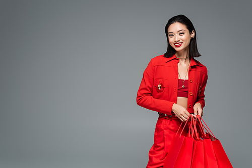 fashionable asian woman smiling at camera while standing with red shopping bags isolated on grey