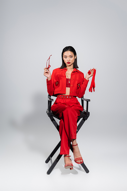 full length of elegant asian woman in red attire holding glove and sunglasses while sitting on grey background