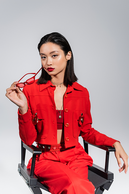 brunette asian woman in red attire sitting on chair with trendy sunglasses isolated on grey