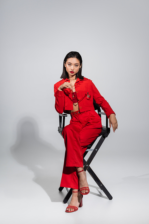 full length of asian model in red jacket and pants holding stylish sunglasses while sitting on chair on grey background