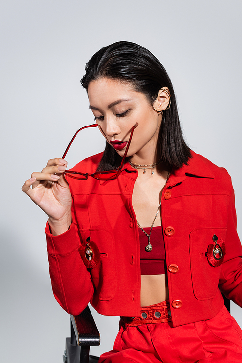brunette asian woman in red jacket holding fashionable sunglasses isolated on grey