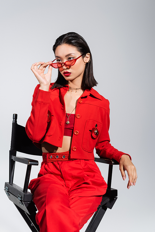 elegant asian woman in red clothes holding sunglasses and looking away while sitting on chair isolated on grey