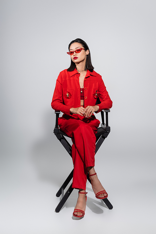 full length of asian model in red elegant outfit and sunglasses sitting on chair on grey background
