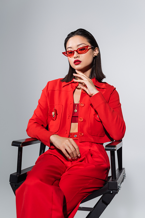 brunette asian woman in red sunglasses and elegant attire sitting on chair and touching chin isolated on grey