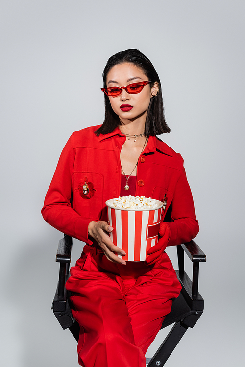 brunette asian woman in red suit and stylish sunglasses sitting with popcorn bucket isolated on grey