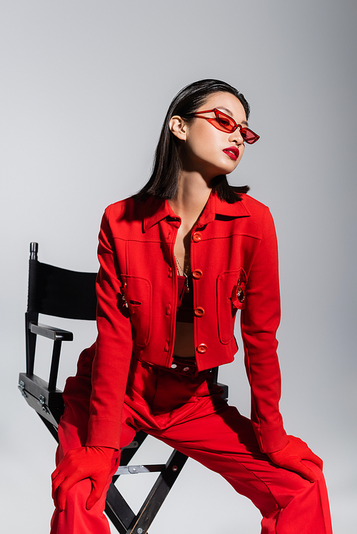 elegant asian model in red jacket and sunglasses posing on chair isolated on grey