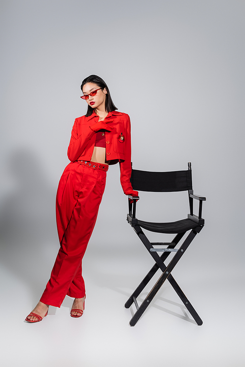 full length of brunette asian woman in red suit and sunglasses posing near chair on grey background