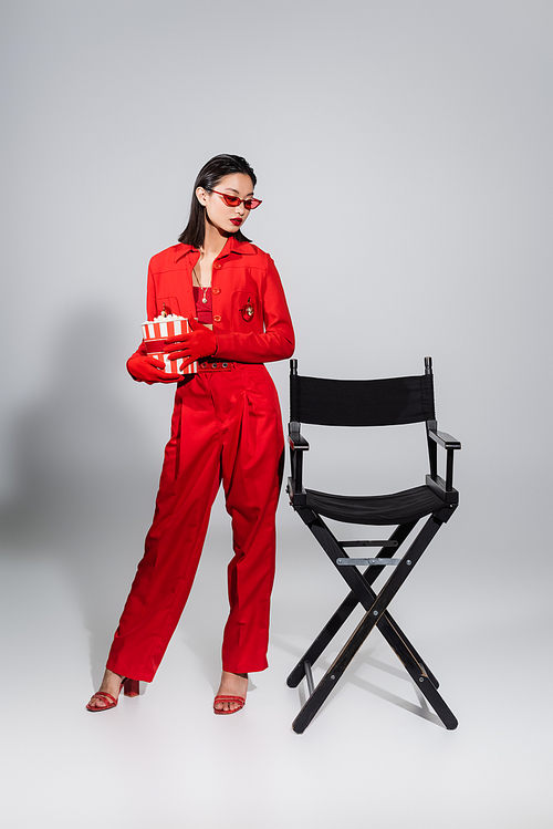 full length of asian woman in red and stylish outfit standing with bucket of popcorn near chair on grey background