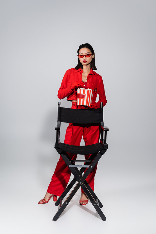 fashionable asian woman in red sunglasses holding bucket of popcorn near chair on grey background