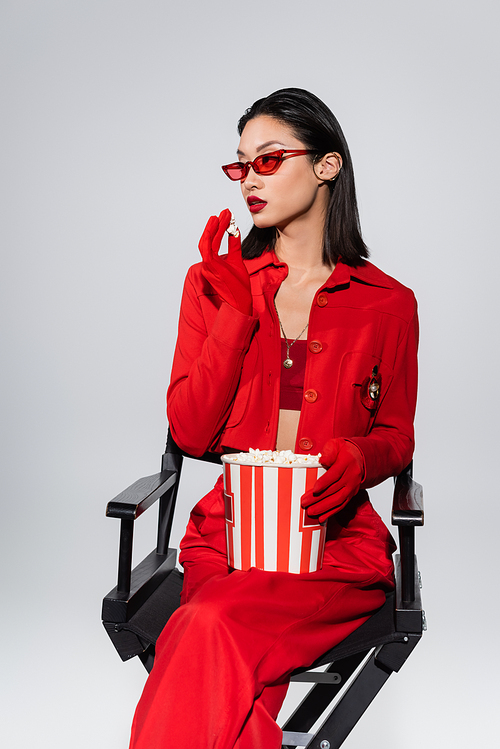 fashionable asian woman in trendy sunglasses and red jacket holding popcorn and looking away isolated on grey