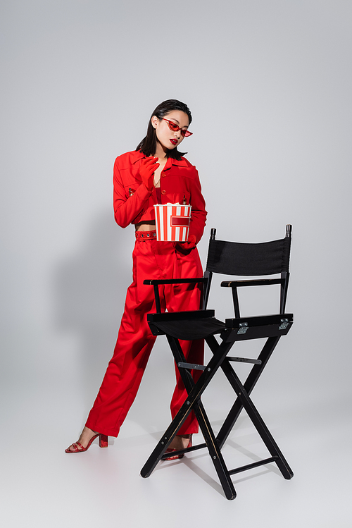 full length of brunette asian woman in trendy sunglasses and red suit standing with popcorn near chair on grey background