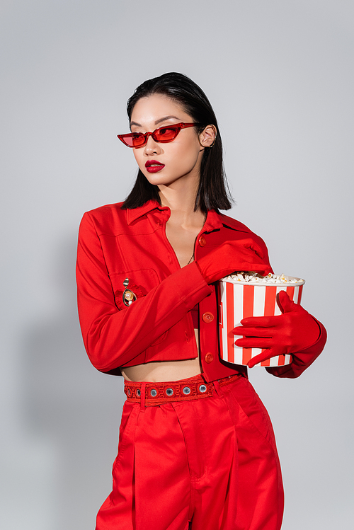 asian woman in trendy sunglasses and red outfit holding bucket of popcorn and looking away on grey background