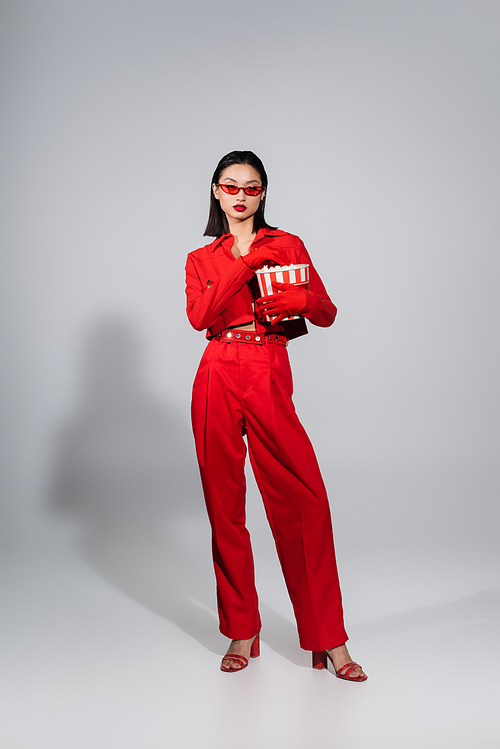 full length of asian woman wearing sunglasses and red suit with gloves holding popcorn bucket on grey background