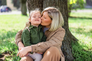 woman kissing happy daughter while sitting under trees in park