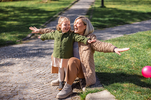 excited mother and daughter with outstretched hands having fun near green lawn in park