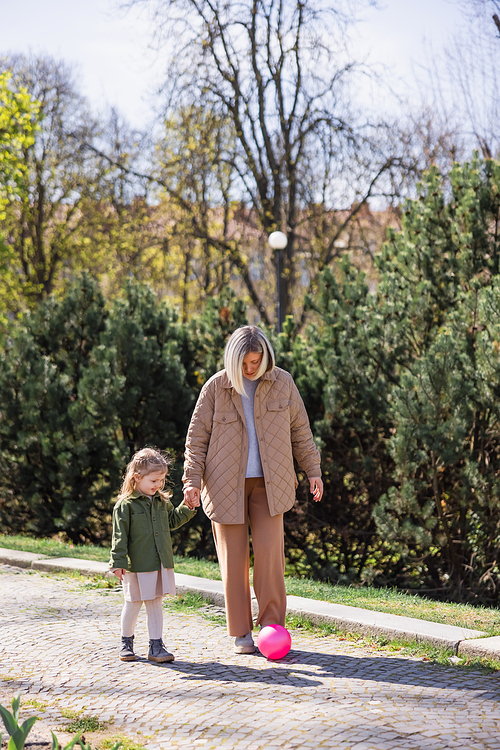 full length view of mother and daughter holding hands while walking near ball in park