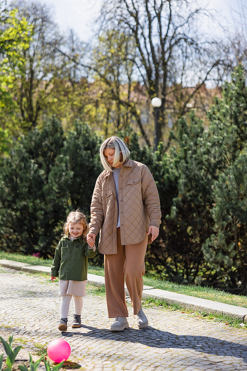 full length view of mother and child in stylish clothes holding hands and walking in park