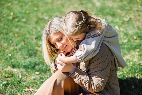 little girl hugging happy mother sitting on green lawn in park