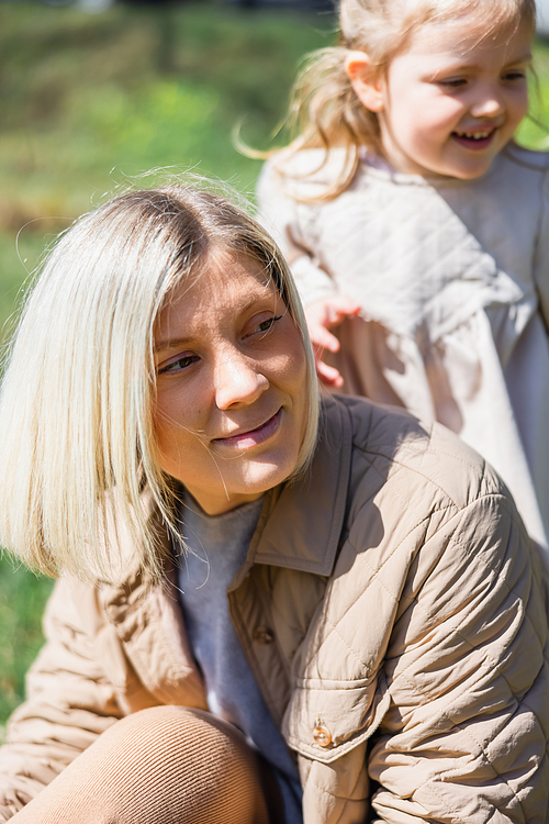 blonde woman smiling and looking away near blurred daughter outdoors