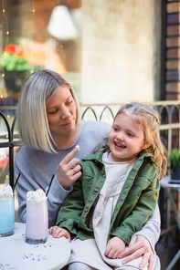 smiling woman with tasty whipped cream on finger sitting with daughter in cafe on street