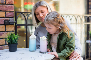 little girl drinking sweet milkshake near mother and flowerpot in cafe outdoors